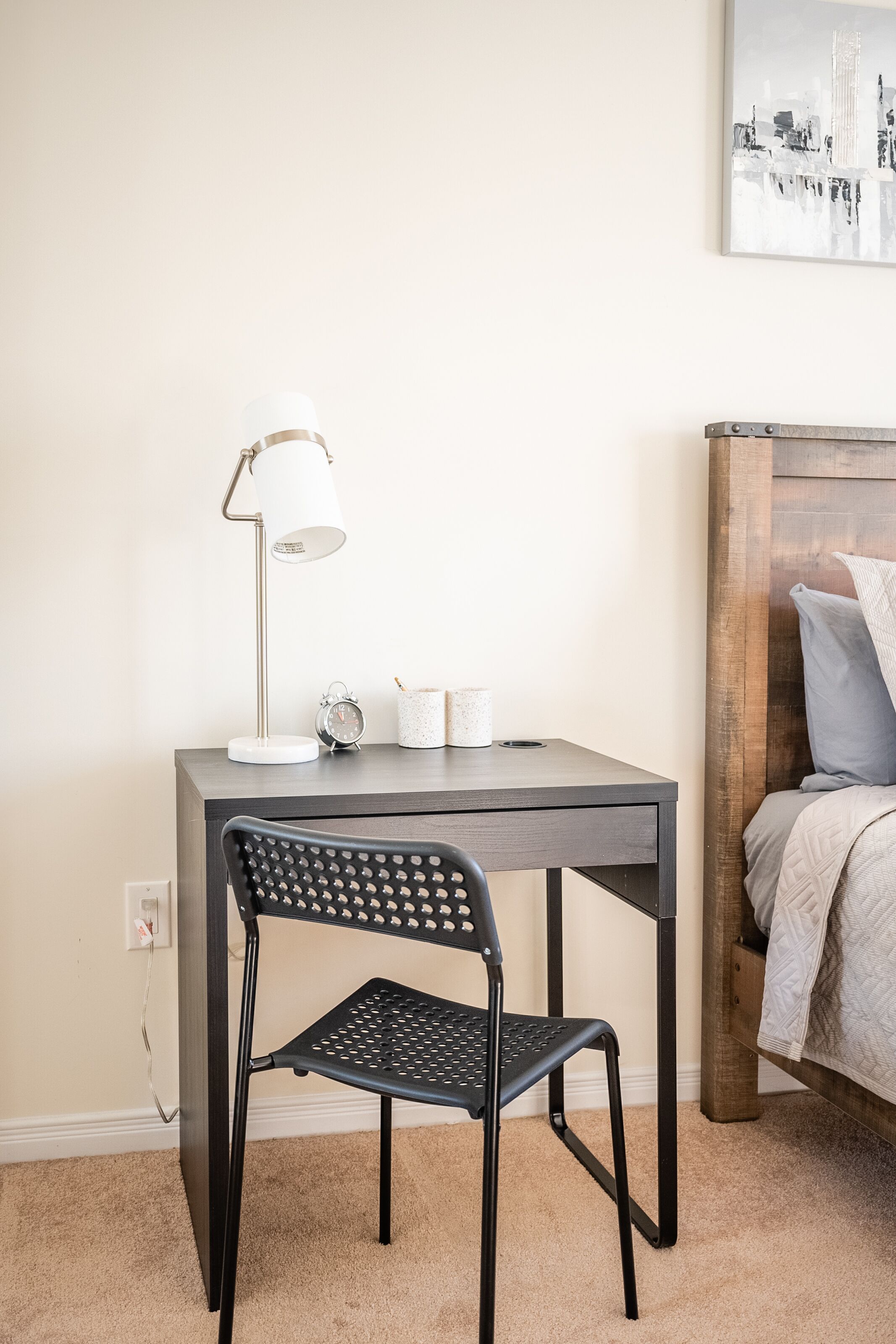 A small, neatly arranged bedroom with a bed, a desk with a chair, a lamp, and a wall-mounted clock.