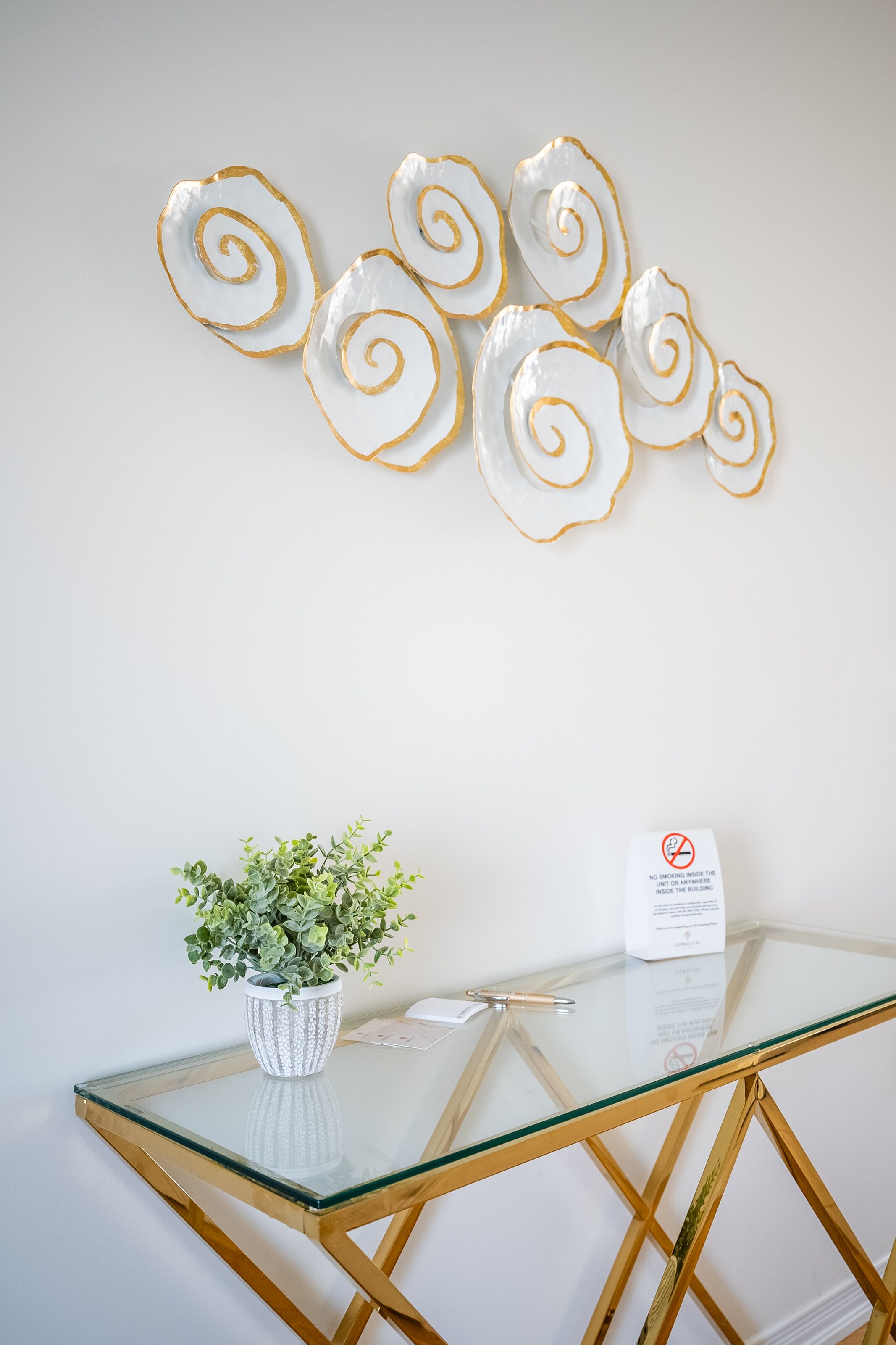 A glass-top table with a white vase and a plant, a wall-mounted decorative piece, and a no-smoking sign.