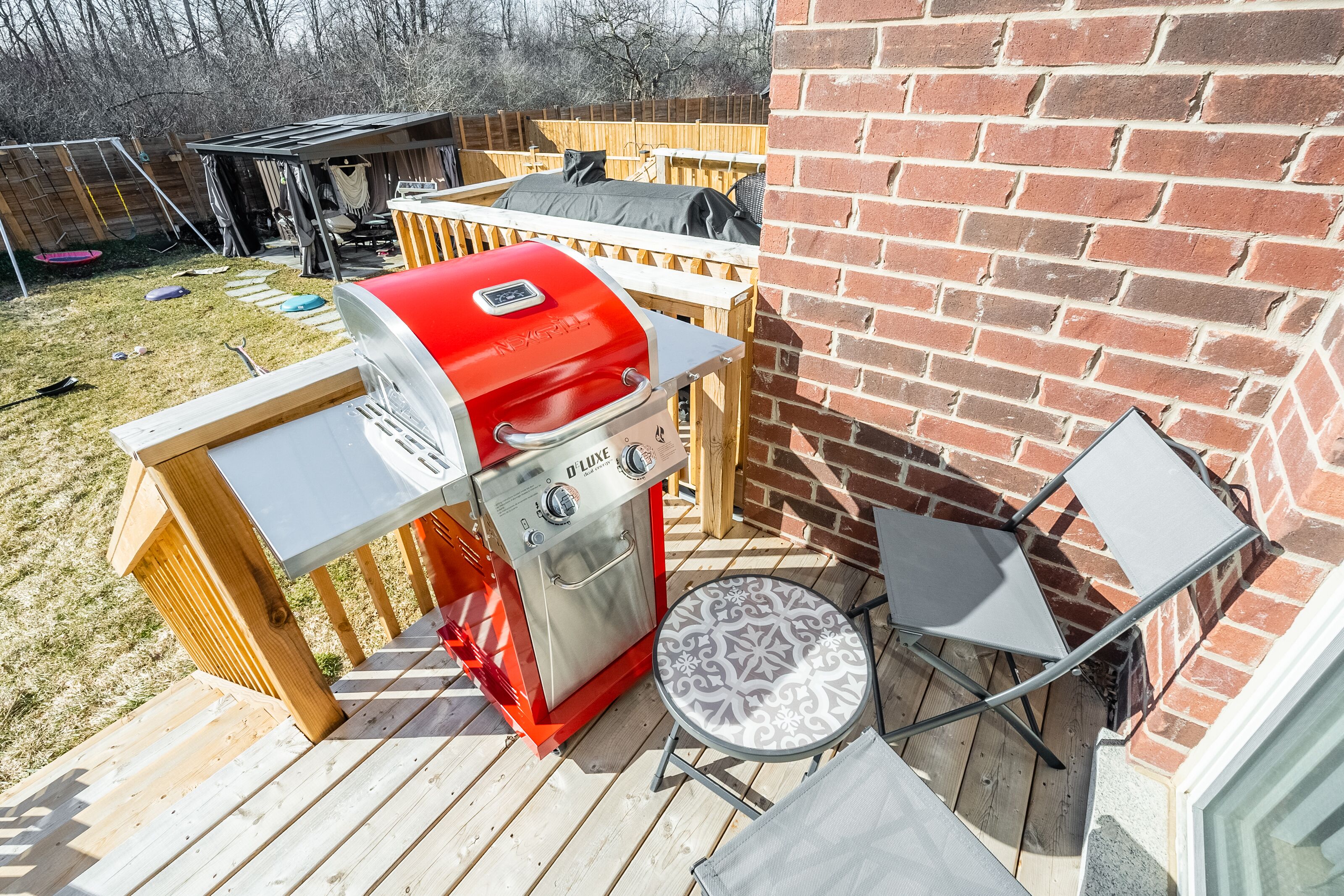 Ein Garten mit einem roten Grill, einer Holzterrasse, einer Ziegelmauer und einem kleinen Patio-Tisch.
