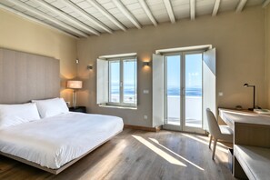 A modern bedroom with a large bed, a desk, and a view of the sea through open windows.