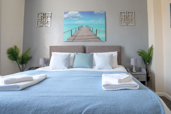 A neatly made bed with a blue blanket and white pillows, a wooden headboard, and a painting of a pier on the wall.
