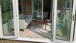 Una habitación moderna con puerta de cristal corredera que da a un balcón. En el interior, hay un sofá blanco, una mesita de madera y una silla azul.