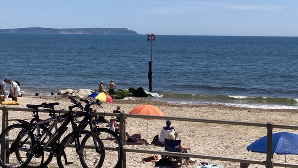 Una playa con bicicletas estacionadas cerca del agua, gente relajándose bajo sombrillas y un puesto de salvavidas al fondo.