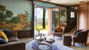 Un salón amplio con un cuadro de un paisaje, un ventanal de vidrio que da a un balcón y una escalera.