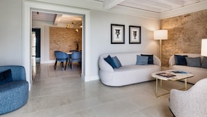 Un salon moderne avec un canapé blanc, des fauteuils bleus et une table basse ronde. On y trouve également un coin repas avec un mur en briques et un lustre.