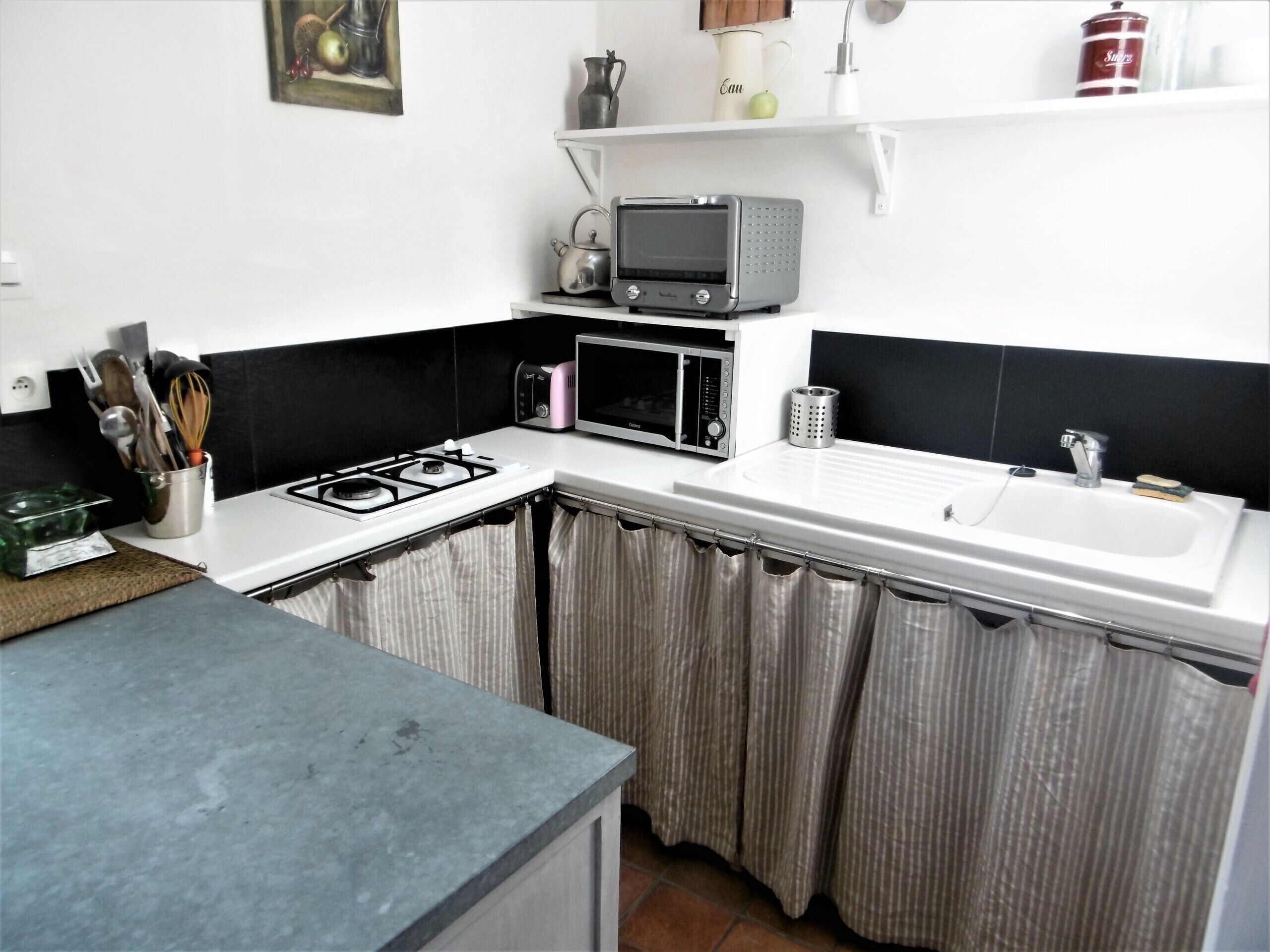 A kitchen with a gas stove, microwave, and sink. There is a shelf above the microwave with a kettle and a container.