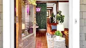 A wooden door leading into a room with a green wall, a brown armchair, a round side table with a plant, and a neon sign. - 1929 Historic Craftsman Bungalow | King Bed (Atlanta)