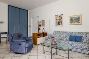 Ein Wohnzimmer mit einem blauen Sofa, einem gemusterten Sofa, einem Glastisch, einem blauen Sessel, einem Schrank und einer Tür.