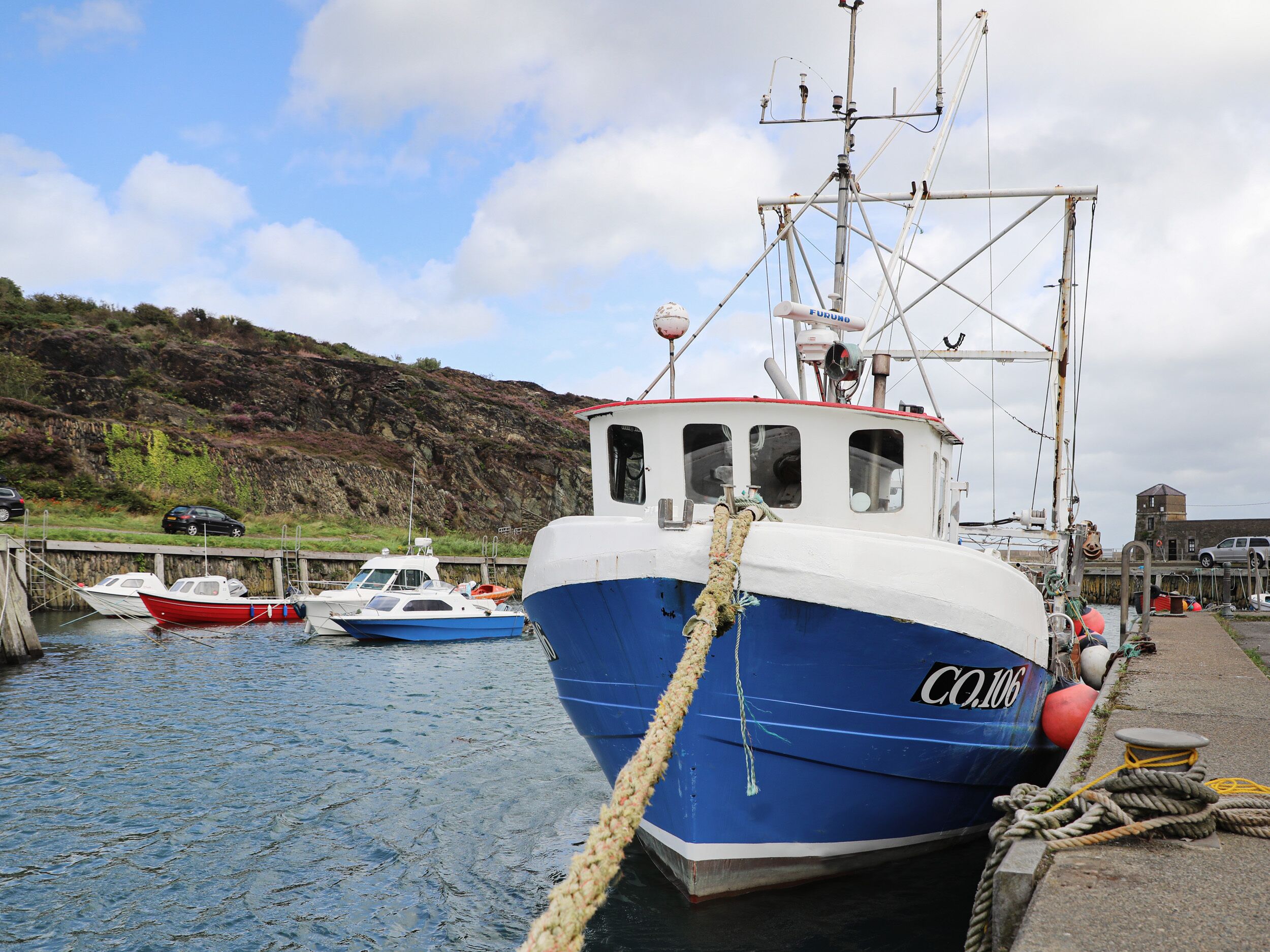 Un puerto con un barco grande azul y blanco amarrado al muelle, rodeado de embarcaciones más pequeñas y una ladera rocosa al fondo.