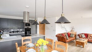Eine moderne Küche mit Edelstahlgeräten, ein Essbereich mit einem Holztisch für vier Personen und ein Wohnzimmer mit Ledersofa und Couchtisch.
