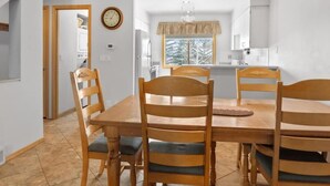 Un coin repas avec une table et des chaises en bois, une horloge au mur et une fenêtre donnant sur les arbres.