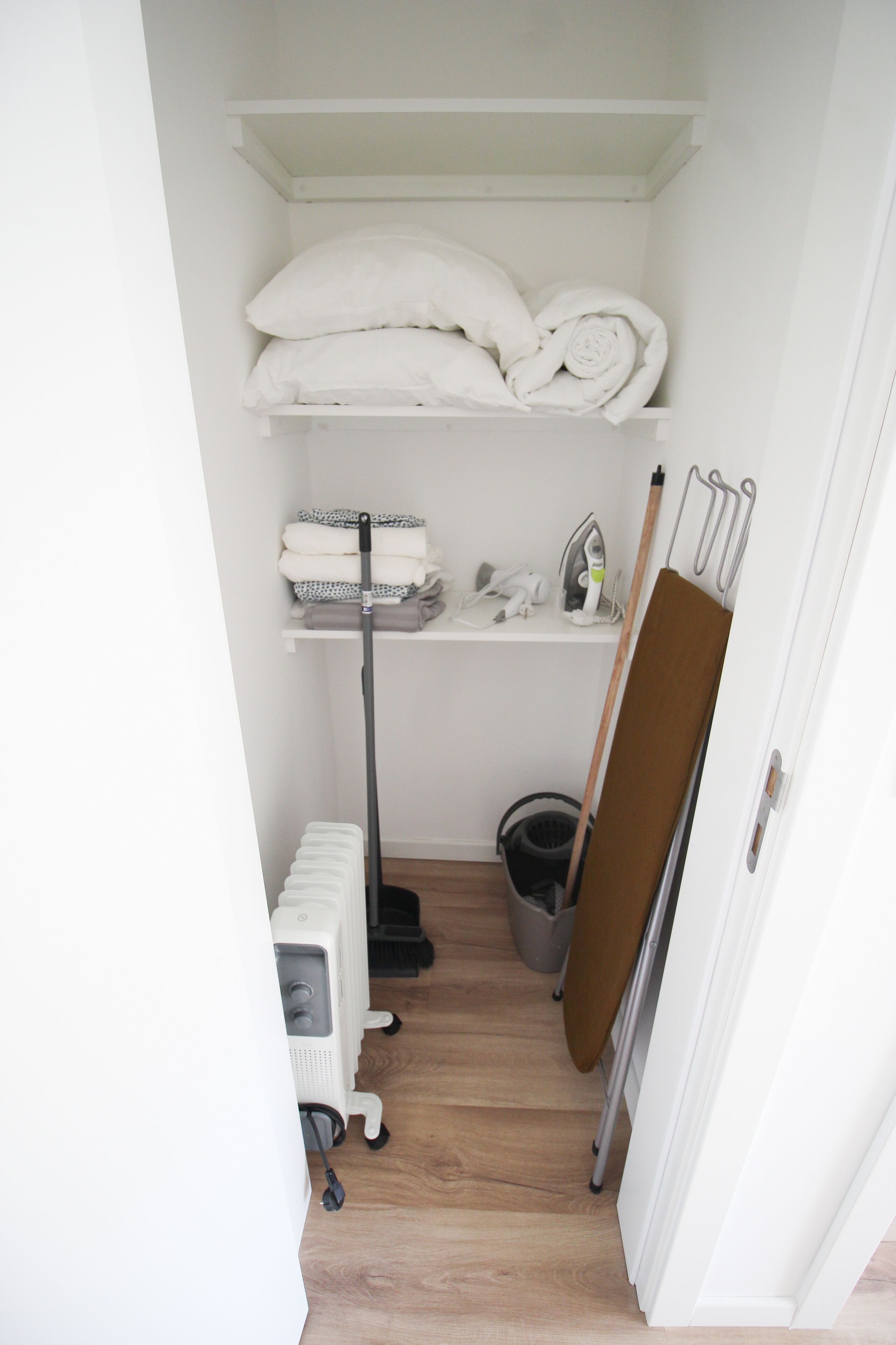A small, clean laundry room with a radiator, ironing board, and shelves with folded towels.