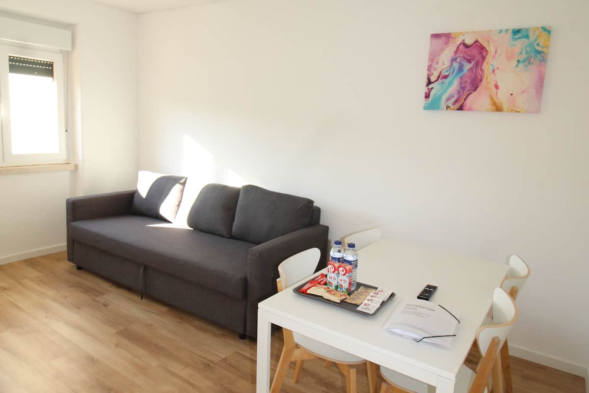 A modern living room with a grey sofa, a white dining table with chairs, a window with blinds, and a colorful abstract painting on the wall.