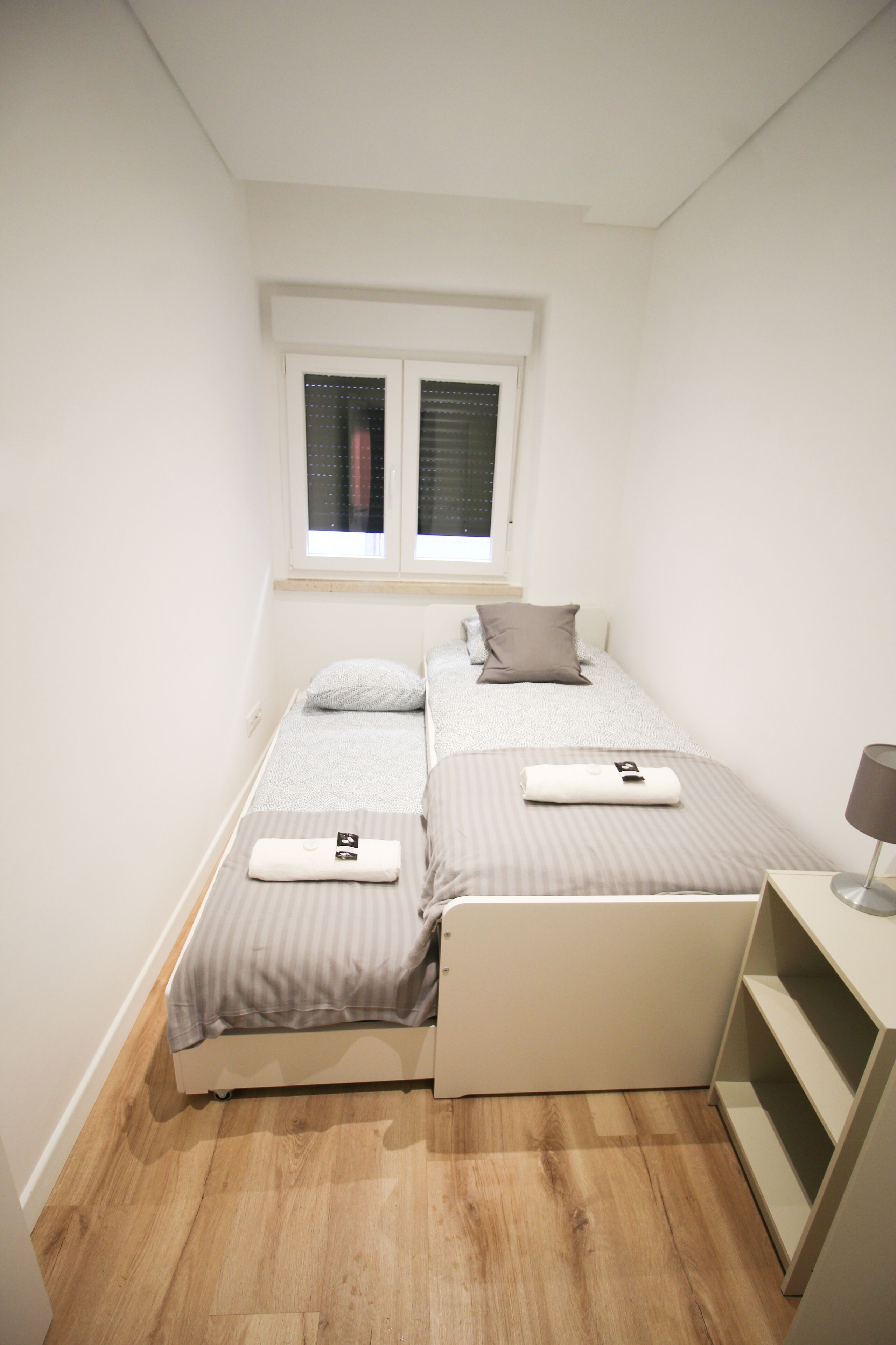 A small, neatly arranged bedroom with two beds, a window with blinds, a bedside table, and a lamp.