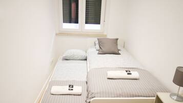 A small, neatly arranged bedroom with two beds, a window with blinds, a bedside table, and a lamp.