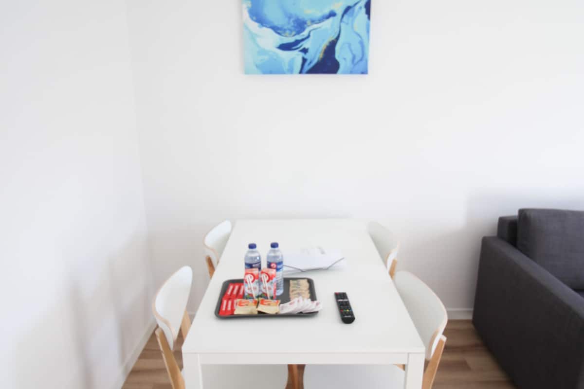 A white dining table with four chairs, a gray sofa, and a blue abstract painting on the wall.