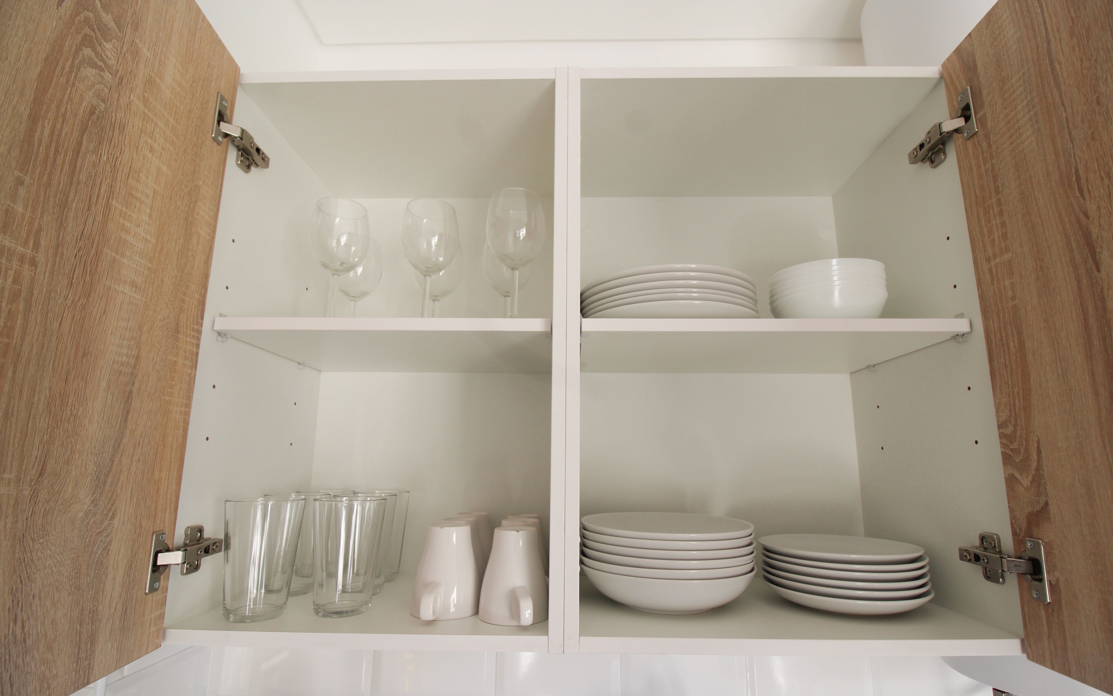 Un mueble blanco con dos estantes; el estante superior contiene copas de vino, y el inferior, platos y tazas.