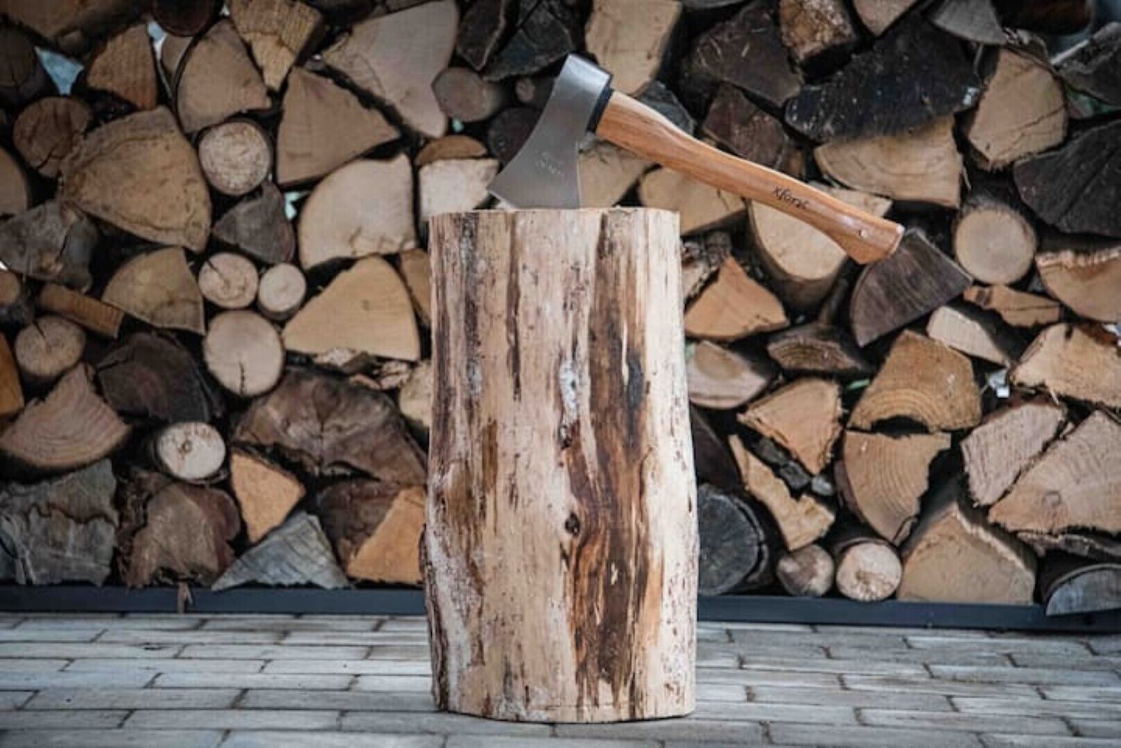 Une hache enfoncée dans une souche d’arbre, avec une pile de bûches en arrière-plan.