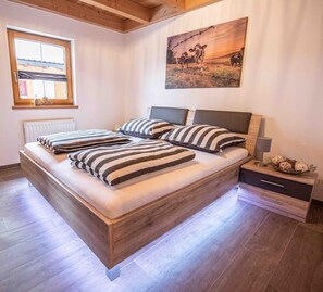 Une chambre à coucher avec un lit en bois, des coussins à rayures, une table de chevet avec une lampe et un tableau représentant des vaches accroché au mur.