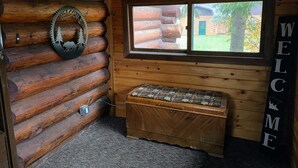 Interno di una baita con panchina in legno, un cartello di benvenuto e una finestra che mostra una casa di legno all'esterno.