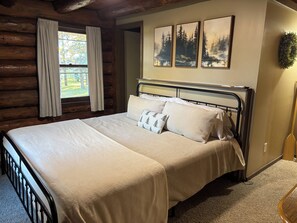 A log cabin bedroom with a metal bed frame, a large window with curtains, and three framed pictures on the wall.