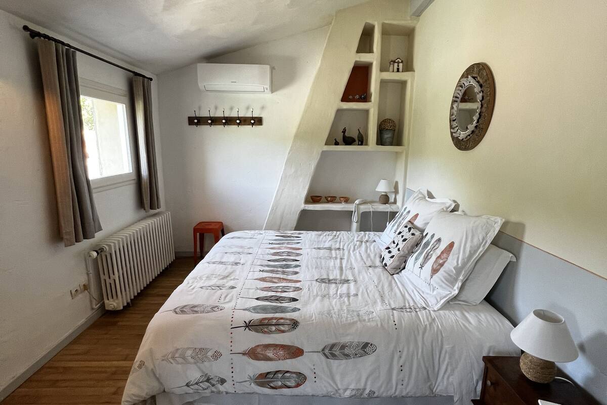 A bedroom with a bed, a nightstand, a lamp, a radiator, a window with curtains, and a shelf with decorative items.