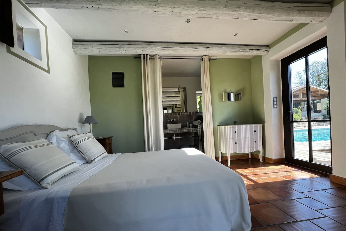 A bedroom with a bed, a nightstand, a bathroom visible through an open door, and a view of a swimming pool outside.
