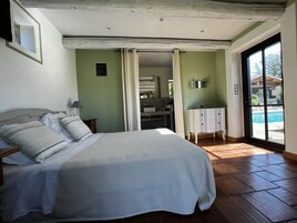 Un dormitorio con cama, mesita de noche, baño visible a través de una puerta abierta y vista a una piscina exterior.