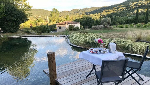 Eine Holzsteg mit einem für eine Mahlzeit gedeckten Tisch, mit Blick auf ruhiges Gewässer und einen üppigen Garten.