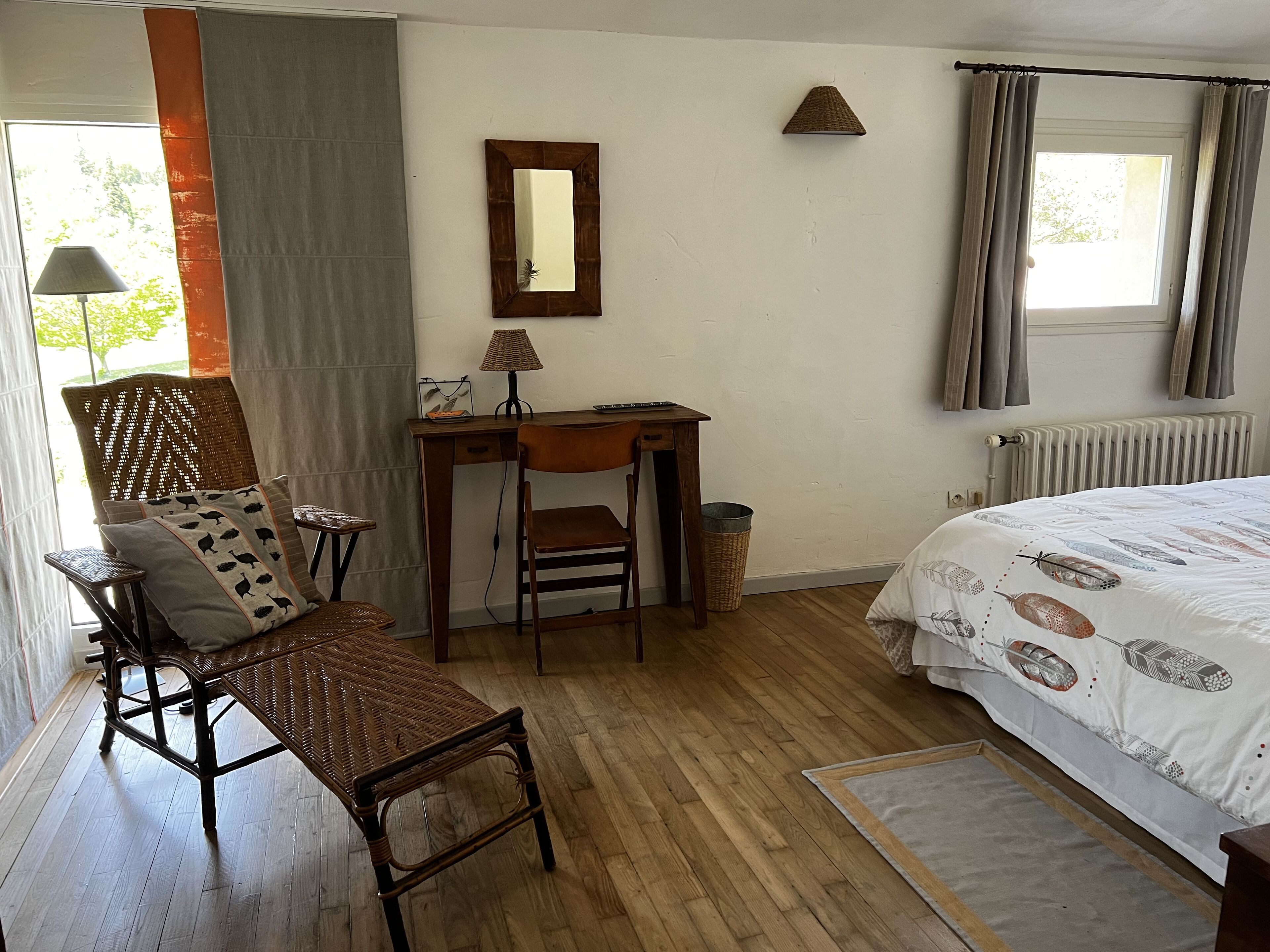 A bedroom with a wooden floor, a bed with a feathered duvet, a desk with a lamp, a chair, and a window with curtains.