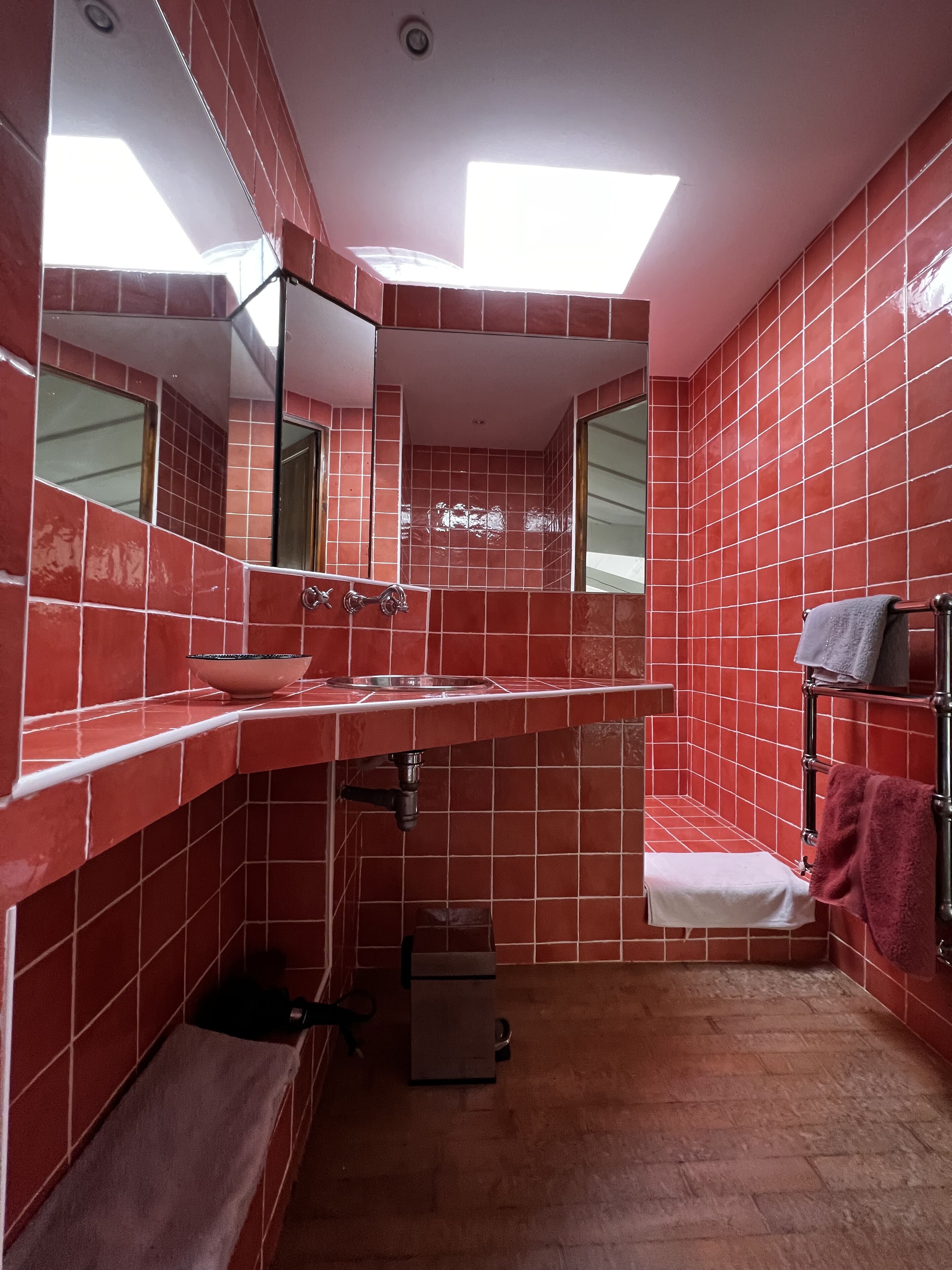 A bathroom with red tiled walls, a sink, a mirror, a towel rack, and a shower area.