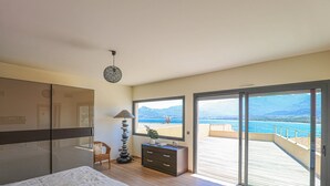 Ein Schlafzimmer mit einem großen Bett, einem Schminktisch, einem Stuhl, einer Lampe und Meerblick durch eine Schiebeglastür.