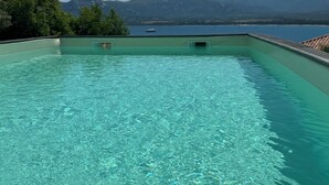 Ein rechteckiger Infinity-Pool mit klarem, blauem Wasser, umgeben von einer niedrigen Mauer und Blick auf einen See sowie Berge im Hintergrund.