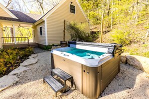 Un jacuzzi con un banco de madera al frente, rodeado por un área de grava y una casa al fondo.