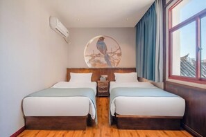 A double room with two beds, a wooden headboard, a wall-mounted air conditioner, a circular artwork of a bird, and a large window with curtains.
