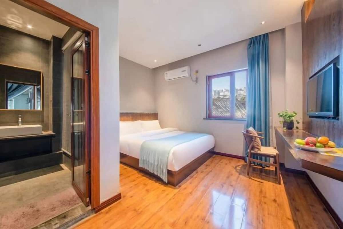 A bedroom with a bed, a chair, a small table with fruits, a window, and an air conditioning unit.