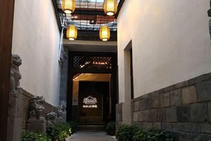 Um corredor estreito com estátuas de leões de pedra à esquerda, que leva a uma entrada de edifício com fachada escura e uma placa sobre a porta.