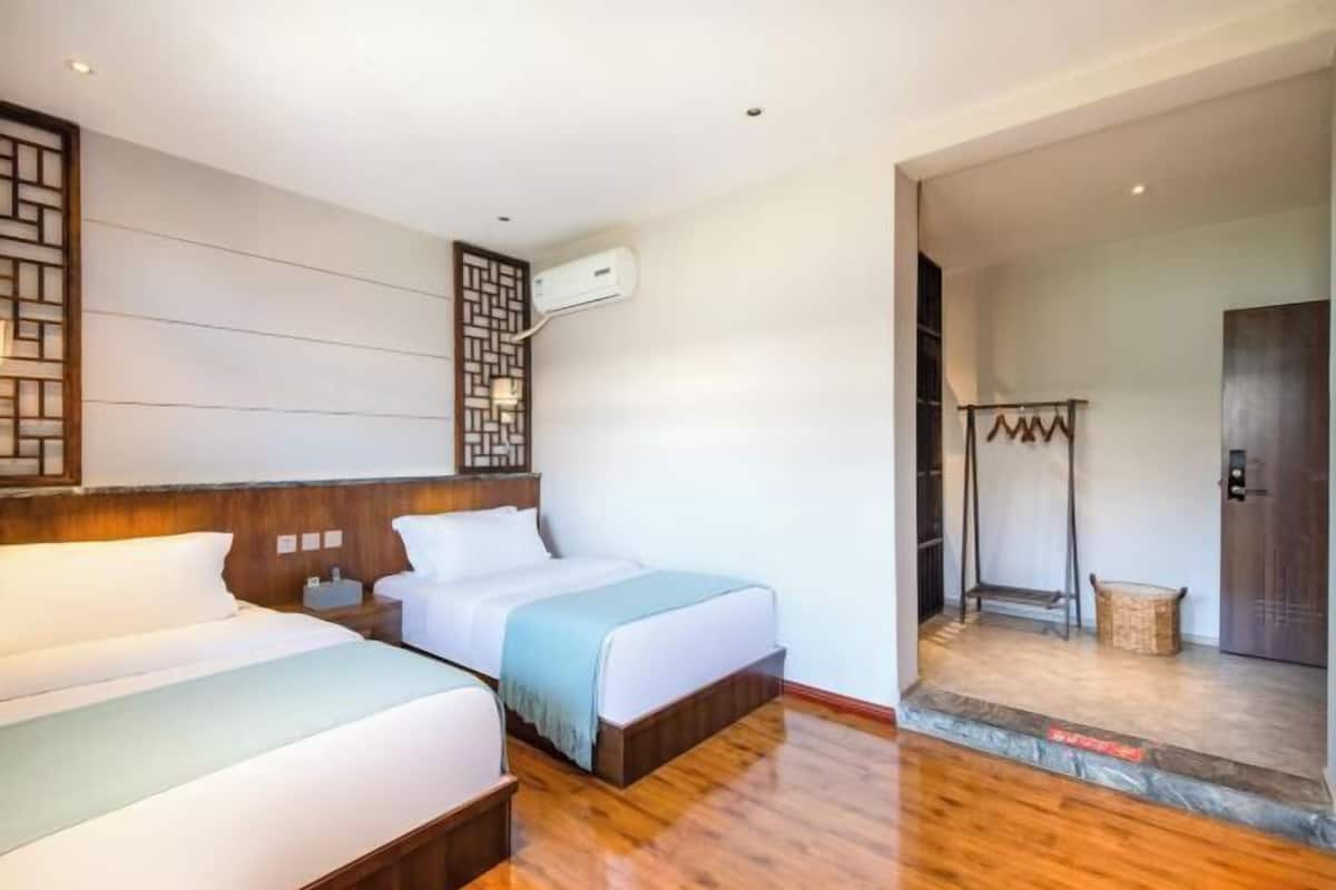 A hotel room with two beds, a wooden headboard, a wall-mounted air conditioner, and a small seating area with a chair and a basket.