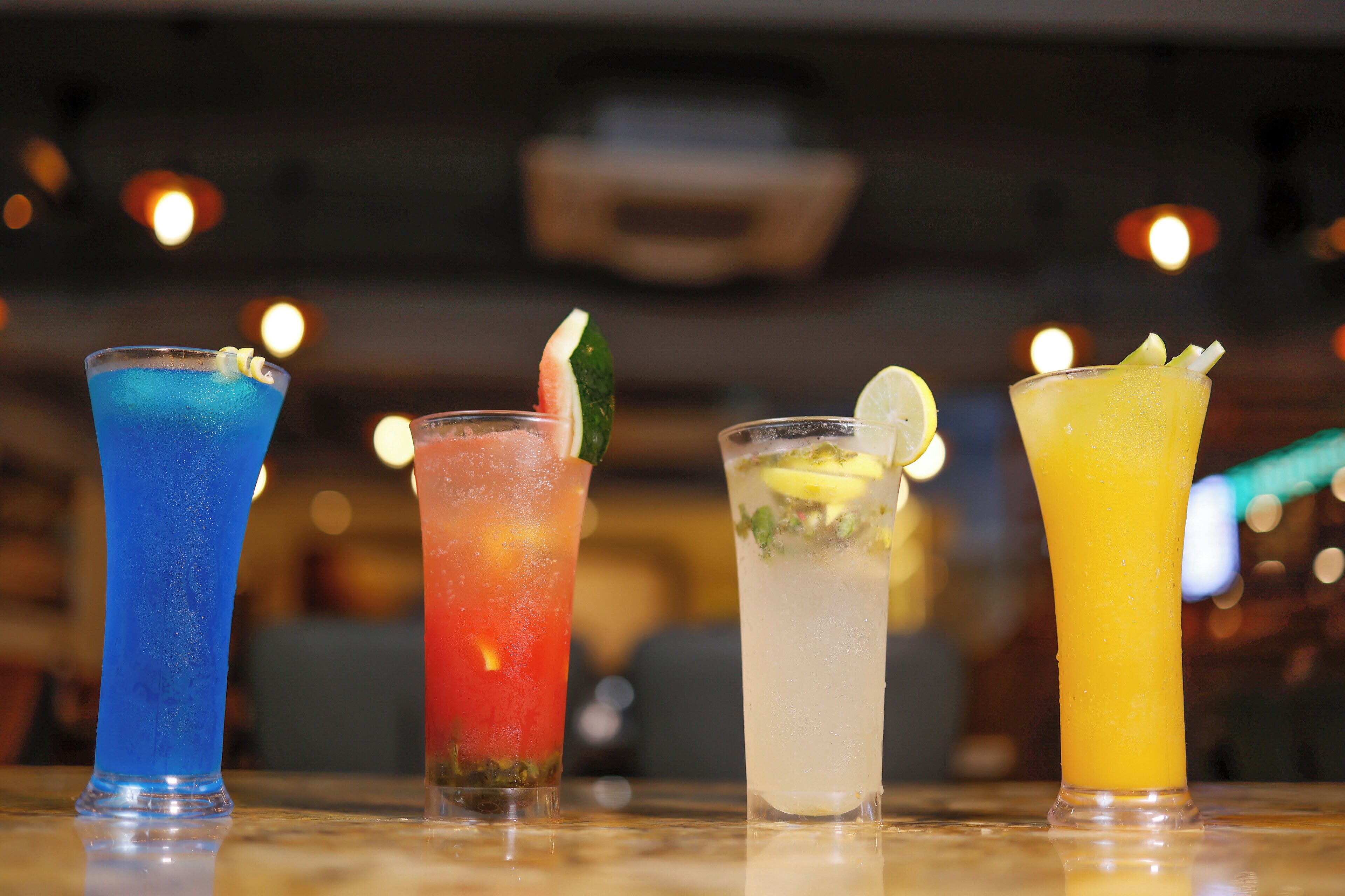 Four cocktails on a bar counter with a blurred background of lights.