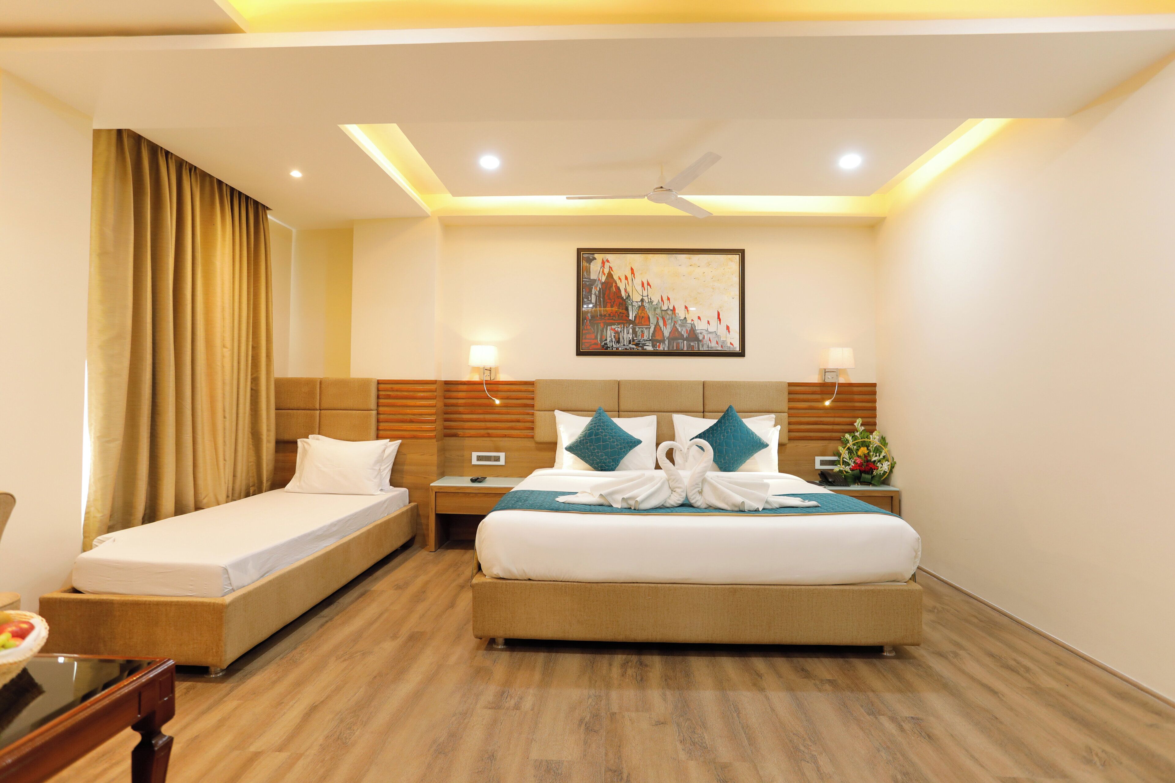 A hotel room with two single beds, a double bed, a desk with a chair, a nightstand with a lamp, and a ceiling fan.