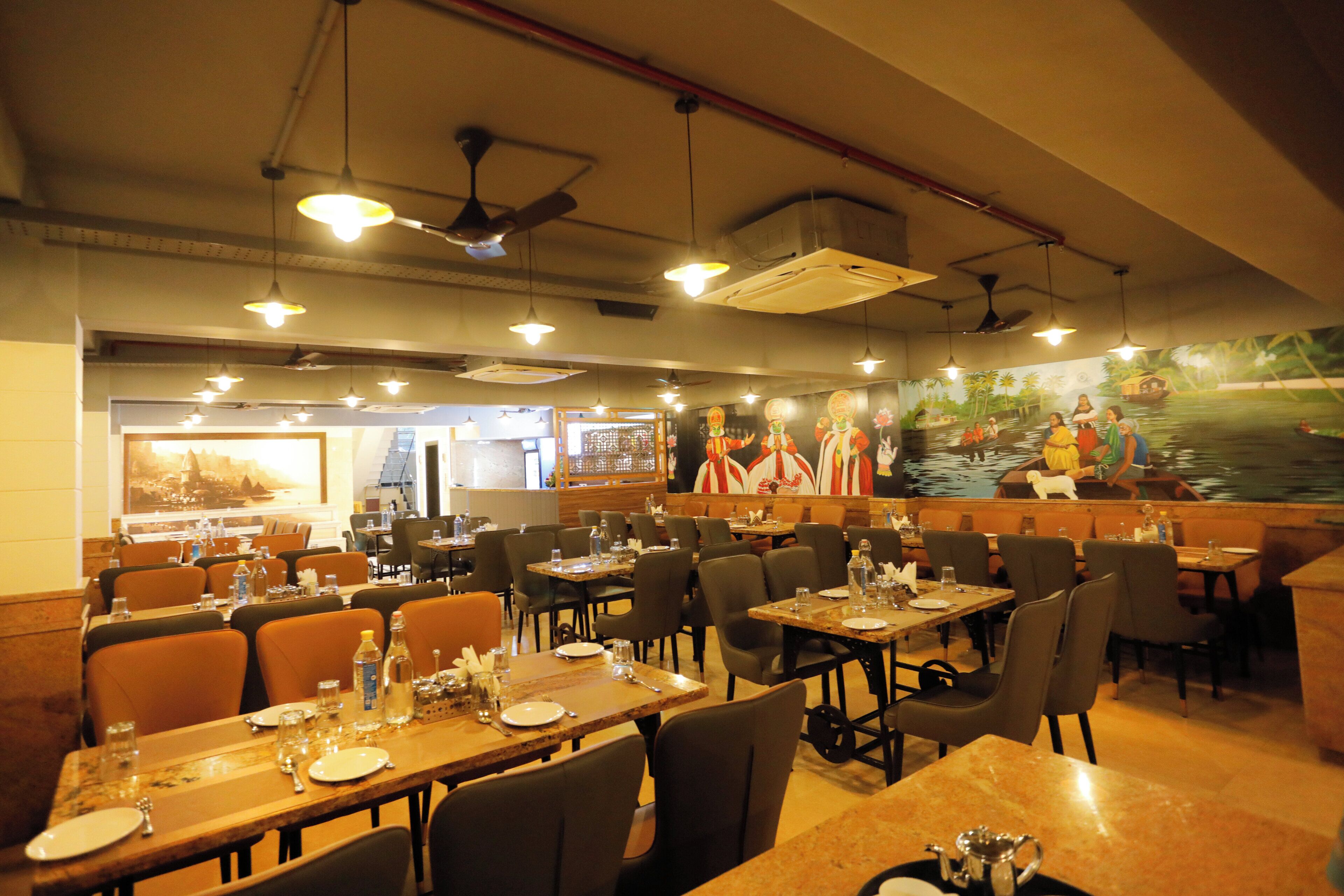 Un restaurant doté de nombreuses tables et chaises, de ventilateurs de plafond et d’une fresque murale représentant un paysage tropical avec des bateaux.