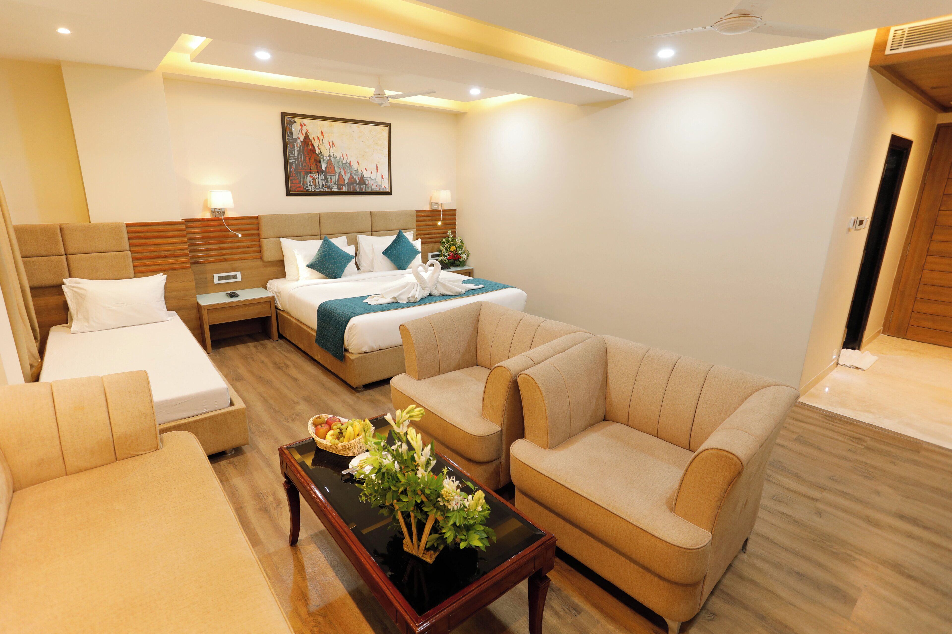 Una habitación de hotel moderna con una cama tamaño king, dos camas individuales, un sofá, una mesa de centro con flores y un cuadro en la pared.