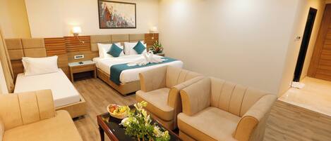 A modern hotel room with a king-sized bed, two single beds, a sofa, a coffee table with flowers, and a painting on the wall.