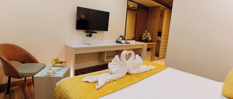 A hotel room with a bed, a desk with a flat-screen TV, a chair, and a small table with a fruit basket and a plate.
