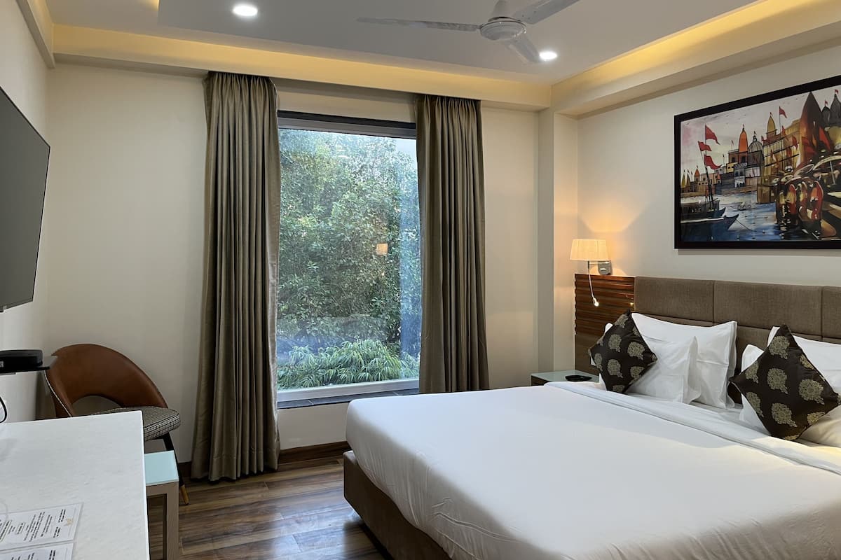 A hotel room with a bed, a desk, a chair, a window with curtains, a ceiling fan, and a painting on the wall.