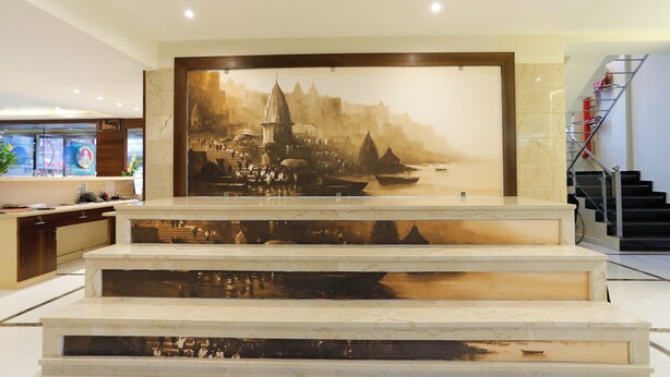 A reception area with a large mural of a historical cityscape, marble steps leading up to a reception desk, and a staircase to the right. - HOTEL DOLPHIN GRAND  (Varanasi)