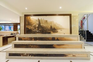 A reception area with a large mural of a historical cityscape, marble steps leading up to a reception desk, and a staircase to the right. - HOTEL DOLPHIN GRAND  (Varanasi)