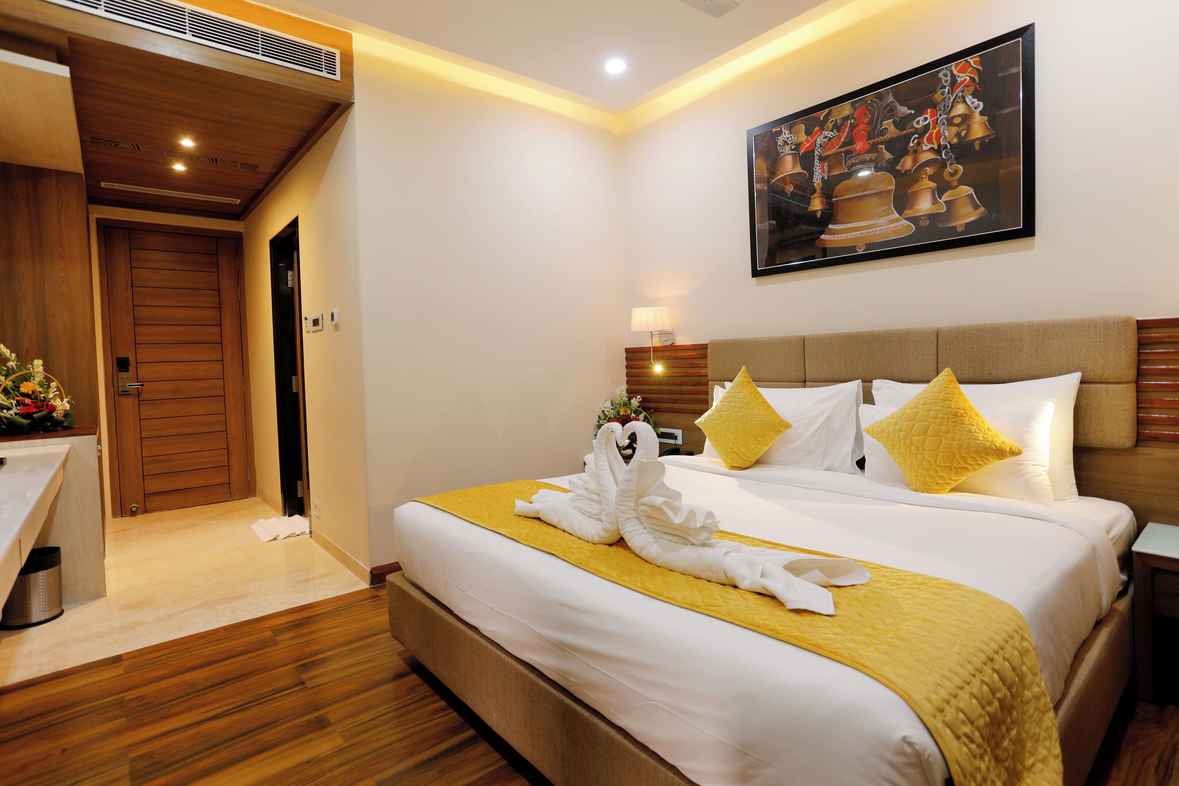 A hotel room with a wooden floor, a bed with white bedding and yellow pillows, a painting on the wall, and a wooden door.