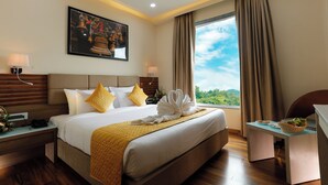 A hotel room with a large bed, a desk, a chair, a window with curtains, a ceiling fan, and artwork on the wall. - HOTEL DOLPHIN GRAND  (Varanasi)