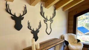 A room with a wooden ceiling, a white wall with three mounted deer antlers, a wooden table with two candles, and a fluffy chair by a window.